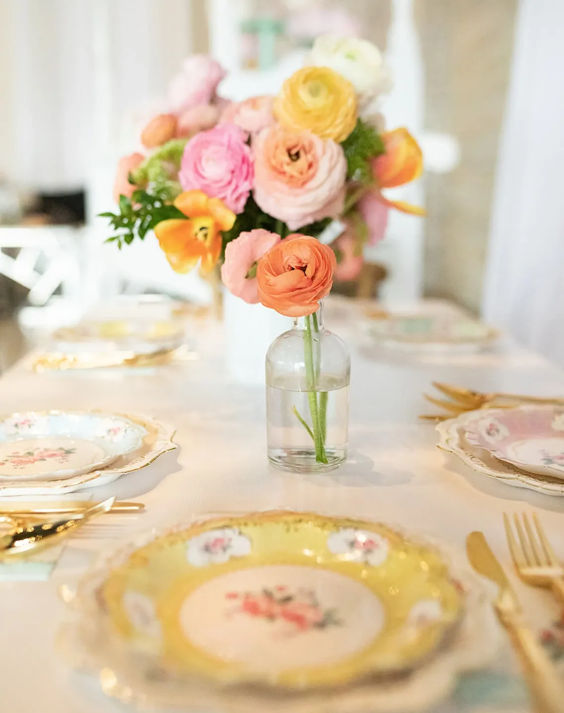 Spring event table with floral china, gold cutlery, and an orange ranunculus in a bud vase