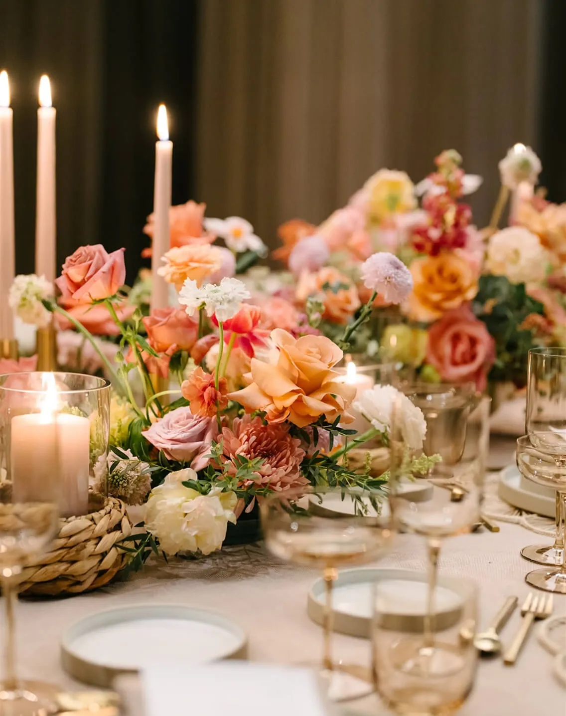Romantic dinner setting with peach floral centerpiece, lit taper candles, and gold flatware