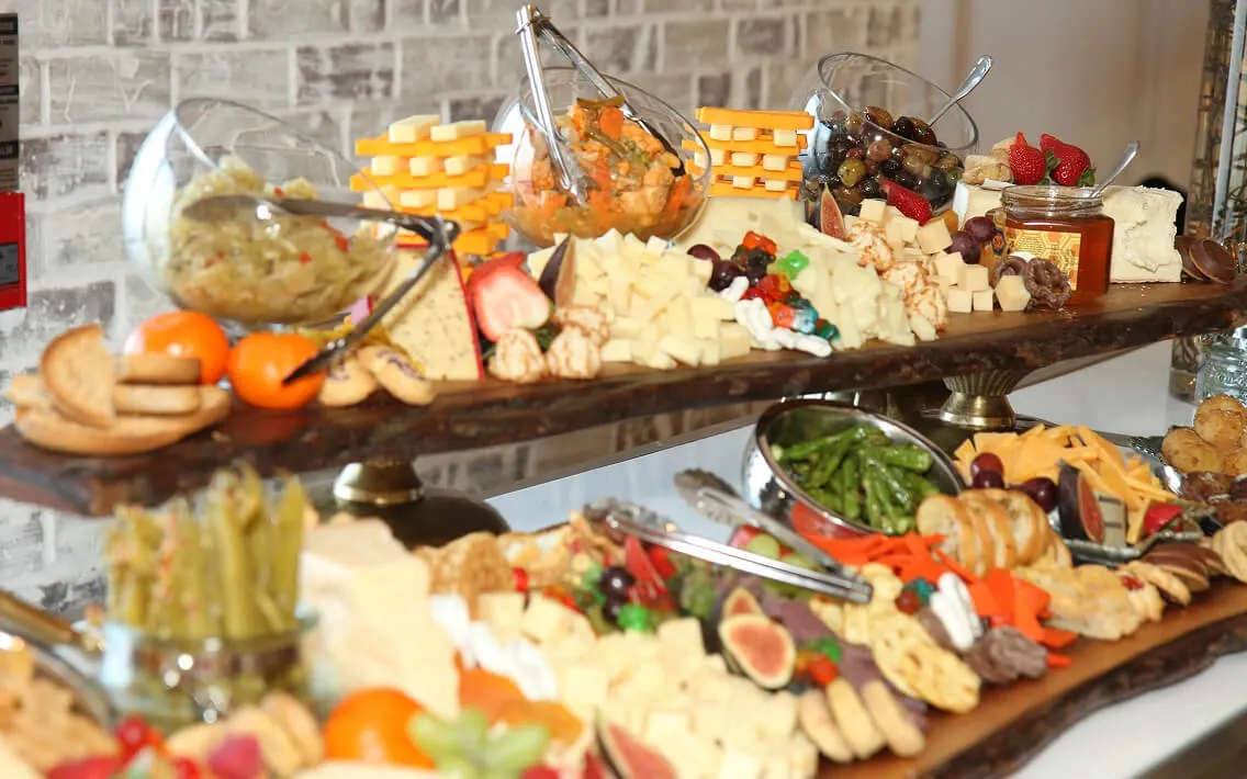 Two-tier wooden grazing display with olives, cheeses, fruit, crackers, and serving tongs