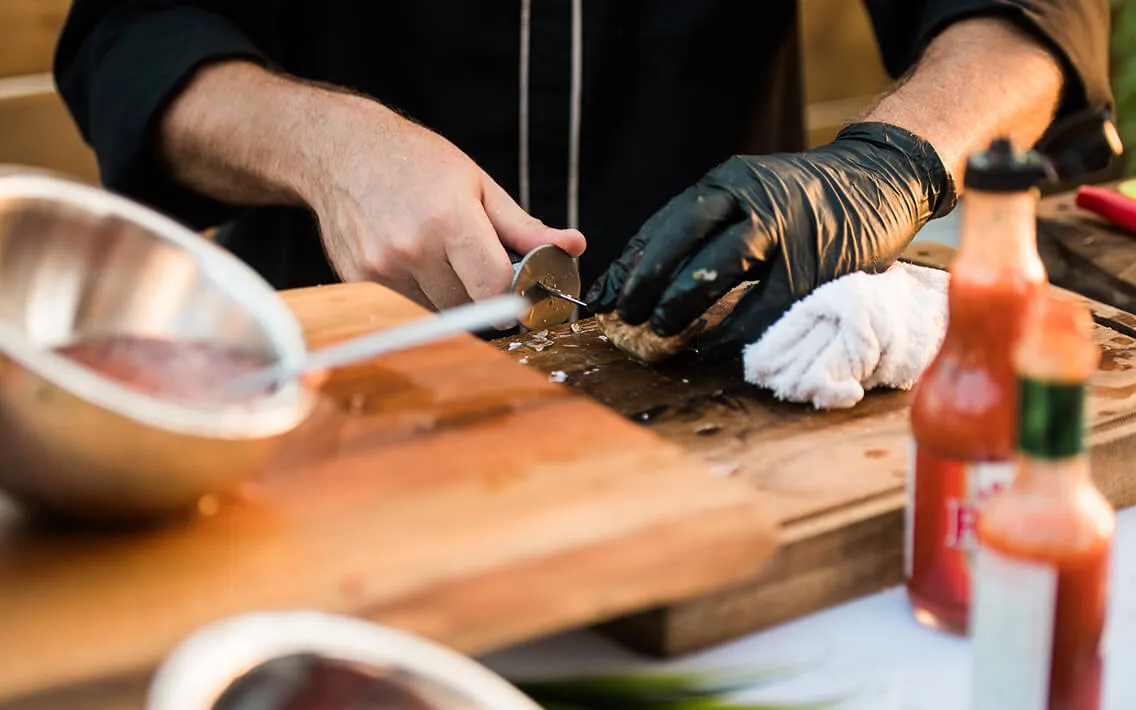 Gloved hands opening fresh oysters next to hot sauce bottles at an event station