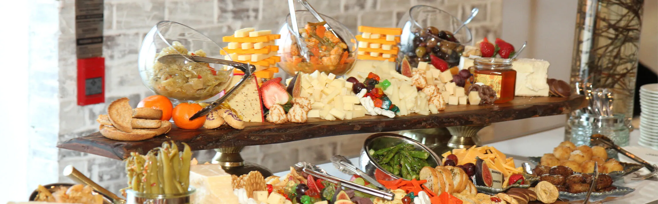Abundant two-tier charcuterie buffet with cheese stacks, olives, and fruit against brick
