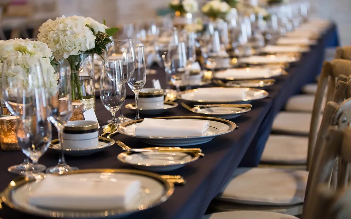 Navy banquet table with gold-rimmed plates, white hydrangeas, and wooden cross-back chairs